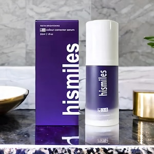 Peut inclure: Une boîte et un flacon violets de sérum correcteur de couleur hismiles V34. La boîte indique "TEETH BRIGHTENING" et "30ml | 1 fl oz". Le flacon et la boîte sont sur une surface réfléchissante.