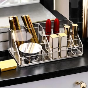 Puede incluir: Organizador de maquillaje de acrílico transparente lleno de varios cosméticos. El organizador contiene barras de labios, brillos de labios, polvos compactos y otros productos de belleza. Las barras de labios son rojas y los brillos de labios son dorados.