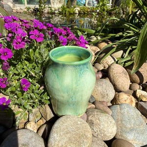 May include: A ceramic vase with a rounded body and a wide opening, featuring a glossy glaze in shades of light green, mint, and turquoise. The surface has subtle variations in colour and texture, with a smooth finish and a slightly darker rim.