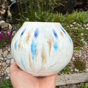 May include: A white ceramic vase with a rounded shape. The vase has blue and brown drip-like patterns. The vase is held in a hand, with a natural outdoor background of plants and water.
