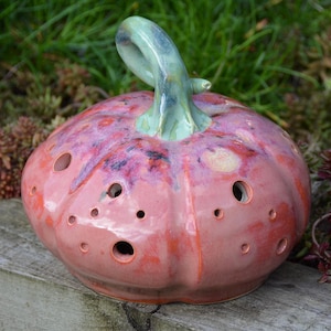 May include: A ceramic pumpkin sculpture featuring a glossy, mottled pink and orange glaze with a smooth, light green stem. The rounded body is punctuated by numerous circular holes of varying sizes distributed across its surface.