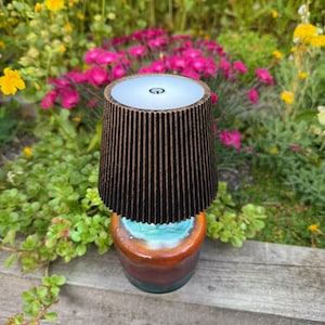 Puede incluir: Una lámpara portátil con una pantalla cilíndrica de color marrón oscuro con nervaduras verticales y una superficie superior blanca con un símbolo de encendido. La base es un recipiente de cerámica redondeado con esmalte brillante turquesa, naranja y marrón.