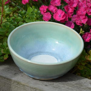 May include: A round ceramic bowl featuring a smooth, glossy glaze that transitions from a soft mint green at the rim to a pale, icy blue toward the centre. The exterior of the bowl displays subtle, horizontal ribbed textures, while the interior base is white.