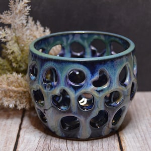 May include: A handcrafted ceramic bowl with a distinctive design. The bowl has a dark blue and teal glaze with numerous circular cutouts. The interior is a darker shade of blue, creating a contrast. The bowl sits on a wooden surface.