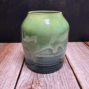 May include: A handcrafted ceramic vase with a gradient of green and blue hues. The vase has a rounded body and a slightly flared rim. The glaze has a glossy finish with a dripped effect, adding texture. The vase is approximately 20 cm tall.