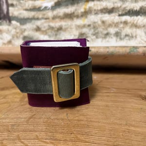 May include: A small, burgundy-colored notebook with white pages, secured by a green leather strap and a gold-colored buckle. The notebook is on a wooden surface, with a painting in the background.