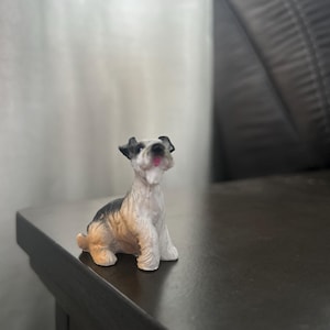 May include: A small figurine of a dog, primarily white with black and tan markings. The dog is sitting upright with its mouth open, tongue visible. The figurine is on a dark brown surface.