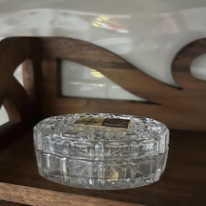 May include: A clear, faceted glass trinket box with a lid, resting on a wooden shelf. The box has a geometric design and a rectangular label on top. The background includes a wooden shelf and decorative elements.