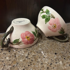 May include: Two ceramic teacups with a floral design. The cups are off-white with pink flowers, green leaves, and a yellow center. The handles are a dark green color.