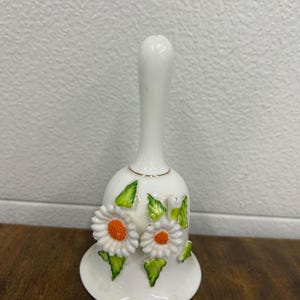 May include: A white ceramic bell featuring a long, slender handle and a flared base. The exterior is decorated with two raised, three-dimensional white daisy flowers with bright orange centers, surrounded by several small, vibrant green painted leaves.