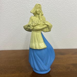 May include: A vintage figurine of a woman in a long blue skirt and a yellow top holding flowers. The figurine has a bonnet and a braided hairstyle. The figure is approximately 5 inches tall.