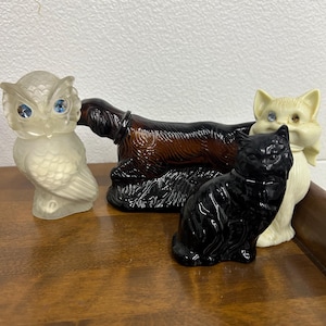 Può includere: Una collezione di statuette di animali vintage, tra cui un gufo, un bassotto e due gatti. Il gufo è bianco sporco con occhi blu, il bassotto è marrone e i gatti sono neri e color crema, ognuno con un occhio di gioiello. Le statuette sono esposte su una superficie di legno.