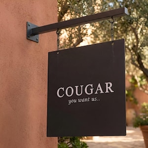 May include: A square black metal hanging sign suspended by two chains from a horizontal black metal arm. The sign features the white text COUGAR in a bold serif font, with the smaller white text you want us.. positioned directly underneath.