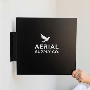 Könnte beinhalten: Ein schwarzes, quadratisches Schild mit dem weißen Text "AERIAL SUPPLY CO." und einer weißen Taubengrafik. Das Schild ist mit einer schwarzen Halterung an der Wand befestigt. Eine Hand hält das Schild.