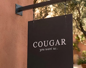 May include: A square black metal hanging sign suspended by two chains from a horizontal black metal arm. The sign features the white text COUGAR in a bold serif font, with the smaller white text you want us.. positioned directly underneath.