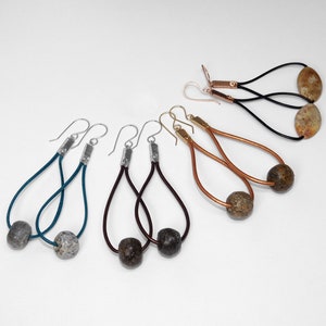 May include: A collection of handcrafted earrings featuring various designs. Some earrings have teal, brown, and black cords with stone beads. Others have copper or gold-toned metal accents and stone or oval-shaped beads. The earrings are displayed on a white background.