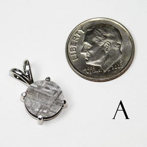 May include: A silver pendant with a round, patterned stone, next to a U.S. dime for size comparison. The pendant has a loop for a chain and is set with four prongs. The stone has a unique, textured appearance.