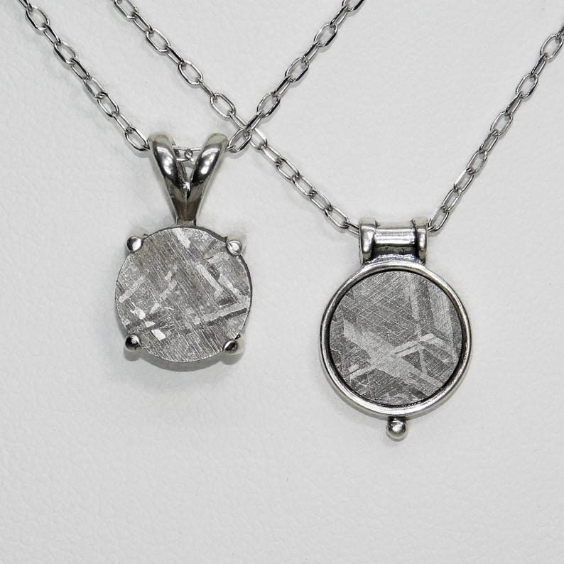 May include: Two silver necklaces with meteorite pendants. One pendant is a round stone held by four prongs. The other pendant is a round stone set in a silver bezel with a small decorative bead at the bottom.