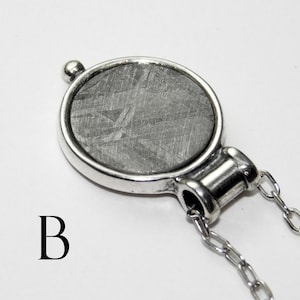 May include: A silver pendant necklace featuring a circular, textured, gray stone set in a silver bezel. The pendant is attached to a delicate silver chain. The letter "B" is printed in black on the lower left.