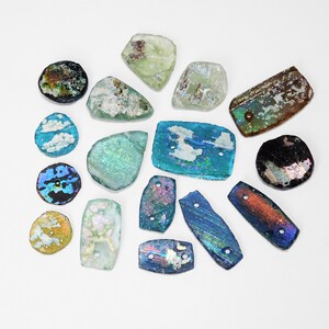 Puede incluir: Una colecci&oacute;n de piezas de vidrio iridiscente de formas irregulares, en varios colores, incluyendo azul, verde y marr&oacute;n. Algunas piezas tienen agujeros, lo que sugiere que son cuentas o componentes para joyer&iacute;a o manualidades. El vidrio tiene un aspecto envejecido.