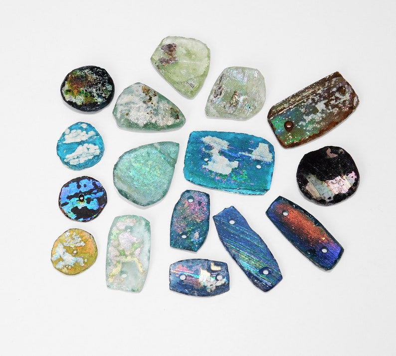 Puede incluir: Una colecci&oacute;n de piezas de vidrio iridiscente de formas irregulares, en varios colores, incluyendo azul, verde y marr&oacute;n. Algunas piezas tienen agujeros, lo que sugiere que son cuentas o componentes para joyer&iacute;a o manualidades. El vidrio tiene un aspecto envejecido.