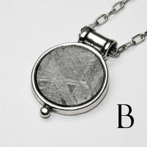 May include: A silver-toned pendant necklace featuring a circular, textured, gray stone set in a silver bezel. The pendant is attached to a delicate chain. The letter "B" is visible in the lower right corner.