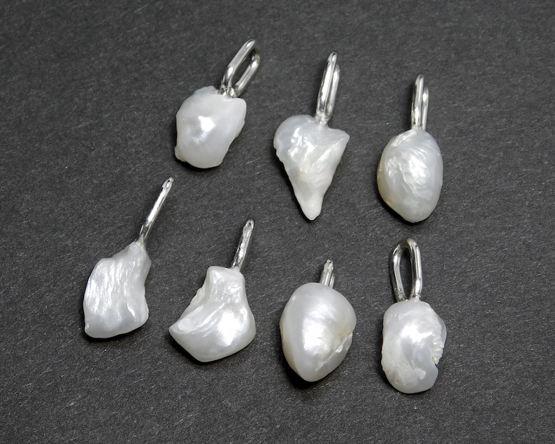 May include: Seven irregularly shaped, white freshwater pearl pendants with silver-colored bails are displayed against a dark gray background. The pearls have a lustrous, organic texture, and each bail is a simple, curved design.