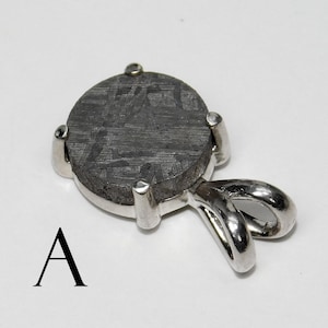 May include: A round, dark gray pendant with a textured surface, set in a silver metal frame. The pendant has a loop for a chain. The letter "A" is printed in black below the pendant.