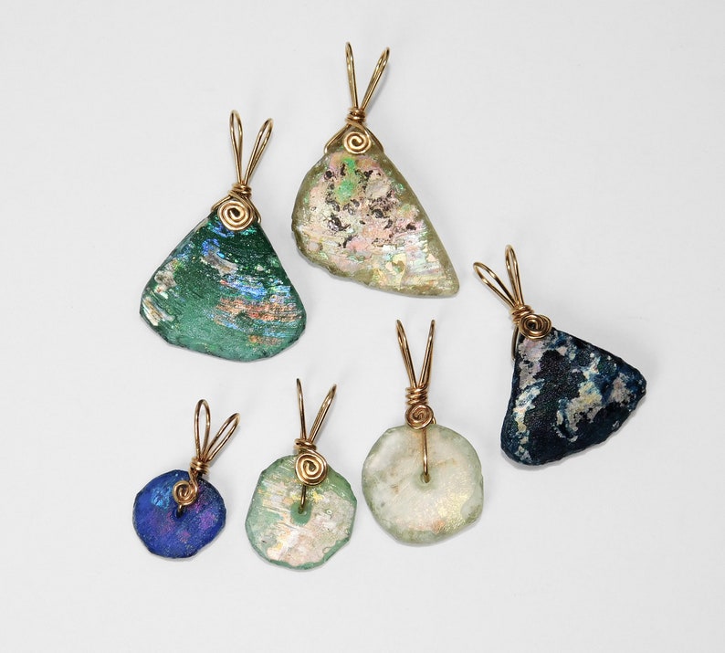 Puede incluir: Colecci&oacute;n de seis colgantes de vidrio marino, cada uno con una forma y color &uacute;nicos, incluyendo verde, azul y transparente. Las piezas de vidrio marino est&aacute;n envueltas con alambre dorado, formando un bucle para una cadena. Los colgantes se muestran sobre un fondo blanco.