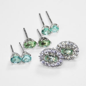 May include: A set of five silver earrings with green and blue gemstones. Two earrings feature a single oval-shaped gemstone, while the other three earrings have a larger oval-shaped gemstone surrounded by a halo of smaller round gemstones.