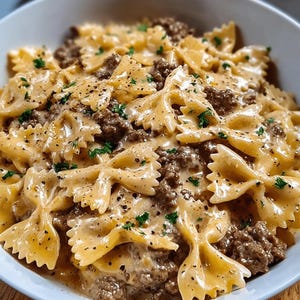 Pode incluir: Uma tigela com massa farfalle cozida, carne moída e um molho cremoso marrom claro. A massa é coberta com salsa fresca e pimenta preta. O prato é servido em uma tigela branca.