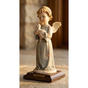 May include: A porcelain figurine of a young angel with curly hair and wings, wearing a long, muted blue robe with a patch detail. The angel holds a small white lamb in its arms. The figure stands on a rectangular wooden base with a gold plaque.