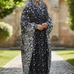 Può includere: Un'abaya nera lunga fino ai piedi con un motivo floreale bianco su tutto il capo, inclusa una copertura per la testa abbinata. L'indumento presenta maniche lunghe e ampie con polsini aderenti e una silhouette fluida, abbinata a scarpe da ginnastica bianche.