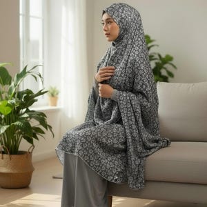 Può includere: Un completo abito da preghiera grigio a fantasia floreale, comprensivo di copricapo e pantaloni coordinati. L'abito è indossato da una persona seduta su un divano beige. La stanza ha una finestra e piante da interno.