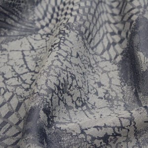 May include: Close-up of a textured fabric with a pattern of overlapping geometric shapes in shades of grey and blue. The fabric has a wrinkled appearance, with light and shadow creating depth.