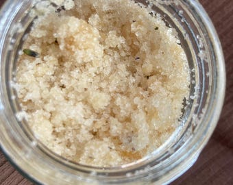 May include: A clear glass jar filled with a light brown, granular substance. The contents appear to be a body scrub or similar cosmetic product. The jar is open, and the scrub fills the container. The background is a solid brown color.