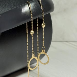 Op de afbeelding: Goudkleurige oorhangers. Elke oorbel heeft een delicate ketting met een klein kralenaccent, een cirkelvormige bedel en een rechte staaf. De oorbellen worden gepresenteerd tegen een zwarte achtergrond.