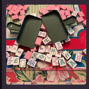 May include: A set of mahjong tiles with white faces featuring pink, green, and blue floral and numerical designs, scattered alongside two olive green plastic trays and a row of pink-backed tiles on a floral patterned game mat.