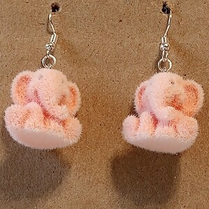 Op de afbeelding: Een paar roze, fuzzy olifant oorbellen met zilverkleurige haken. De oorbellen zijn lichtroze van kleur en hebben een zachte, pluche textuur. De olifanten zitten.