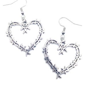 Puede incluir: Un par de pendientes plateados en forma de corazón. Cada pendiente presenta un contorno de corazón con un diseño de alambre de púas. Los pendientes tienen un cierre de gancho para orejas perforadas. La joyería se muestra sobre un fondo blanco.