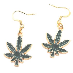 Puede incluir: Un par de pendientes colgantes con dijes de hoja de cannabis en esmalte verde oscuro y bordes de metal dorado. Cada dije cuelga de un gancho de metal dorado, mostrando un diseño detallado de hoja serrada con un acabado brillante.