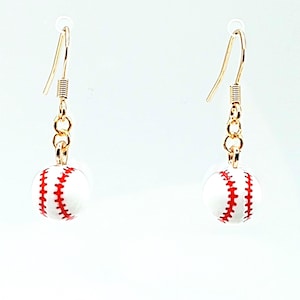 Könnte beinhalten: Ein Paar Baseball-Ohrringe. Jeder Ohrring zeigt einen kleinen, weißen Baseball mit roten Nähten, der an einem goldfarbenen Haken und einer Kette hängt. Die Ohrringe sind vor einem schlichten weißen Hintergrund.