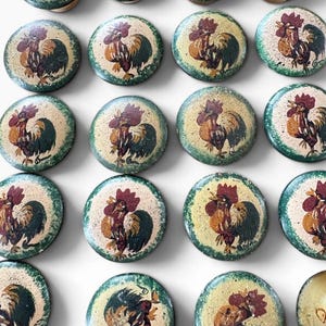 May include: A collection of round wooden cabinet knobs, each featuring a hand-painted rooster design. The roosters are depicted in shades of red, brown, and green against a cream-colored background with a green speckled border.
