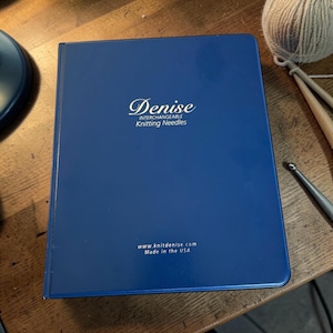 May include: A navy blue case for Denise Interchangeable Knitting Needles. The case has the brand name in a cursive font. The website address and "Made in the USA" are printed at the bottom. A ball of yarn and knitting needles are in the background.