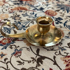 May include: A vintage brass candle holder with a circular base, a central cup for the candle, and a curved handle. The candle holder has a warm, golden color and is likely made of solid brass. The design is simple yet elegant, suitable for various decor styles.