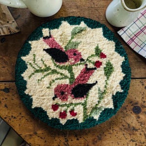 May include: Round, hand-stitched coaster featuring a design of two birds perched on a branch with red berries. The birds have pink bodies and black wings. The coaster has a cream-colored center with a dark green border.