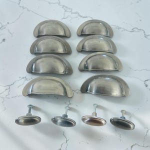 May include: A collection of eight semi-circular brushed nickel cup drawer pulls arranged in two columns, accompanied by four matching oval-shaped cabinet knobs with threaded metal mounting screws extending from the back of each piece.