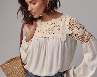 May include: Cream-colored blouse with crochet floral detailing on the neckline and sleeves. The long-sleeved top is paired with high-waisted blue denim jeans. A woven tote bag with brown leather straps is carried over the shoulder.