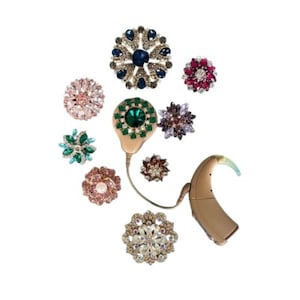 May include: An assortment of bejeweled brooches in various floral designs, featuring a range of colors including pink, green, blue, and clear crystals. A beige hearing aid with a curved design is also present, connected to a small, green-jeweled component.