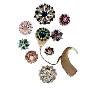 May include: A beige behind-the-ear hearing aid connected to a circular, green-jeweled decorative cover, surrounded by nine assorted floral-shaped brooches featuring various colored crystals, rhinestones, and metallic settings in circular arrangements.